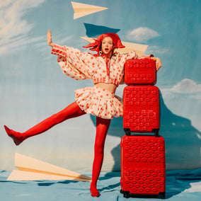 Woman in a polka dot dress and red tights posing with red suitcases against a blue sky.