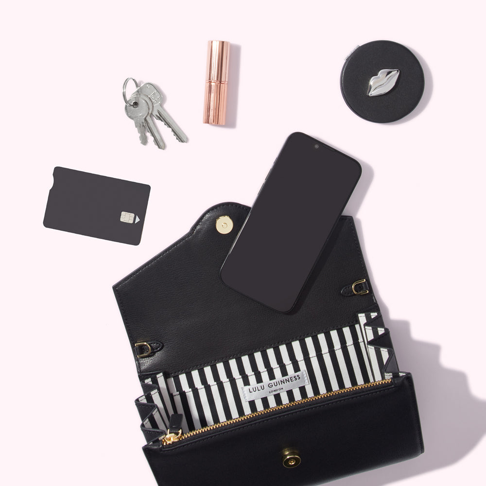 A black Lulu Guinness wallet is open, showcasing its black and white striped interior. A smartphone, keys, a credit card, lipstick, and a compact mirror rest on a white surface nearby.
