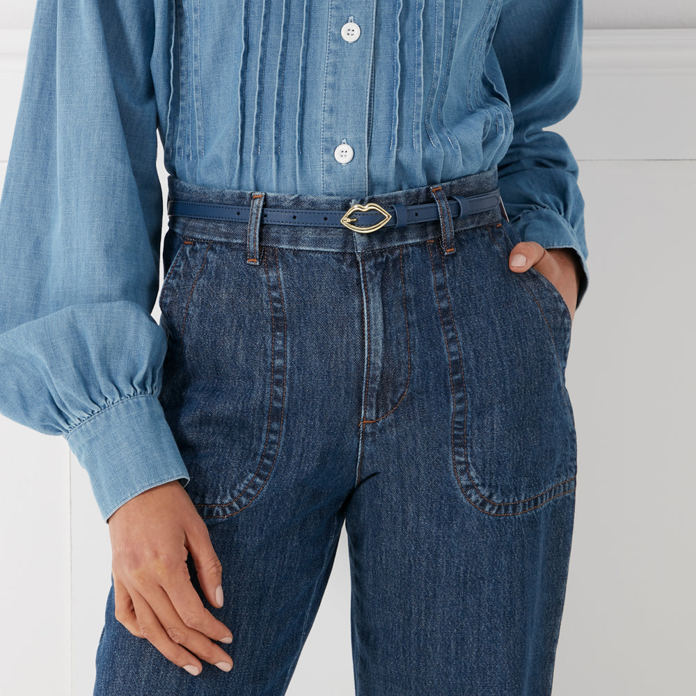 Close-up of a woman wearing a navy blue denim shirt and jeans with a thin navy blue belt featuring a gold lip-shaped buckle.