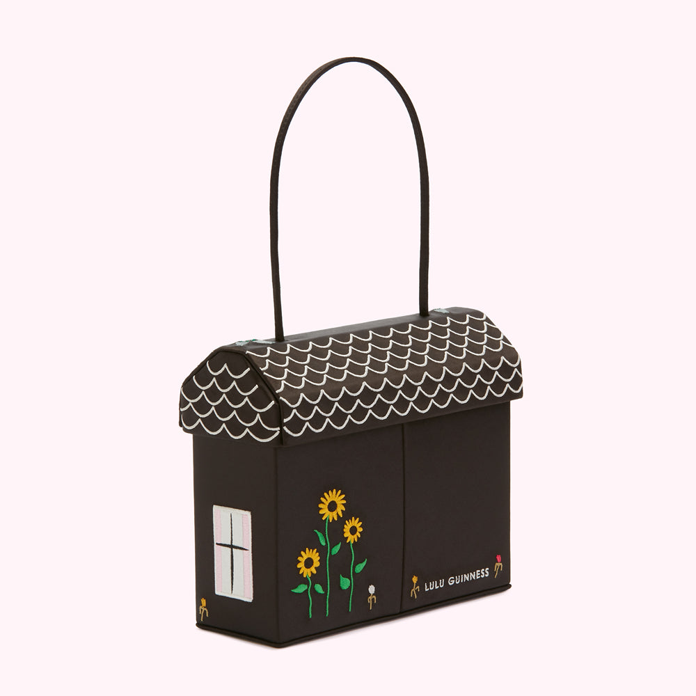 Black house-shaped handbag with a white scalloped roof, sunflowers and the Lulu Guinness logo.
