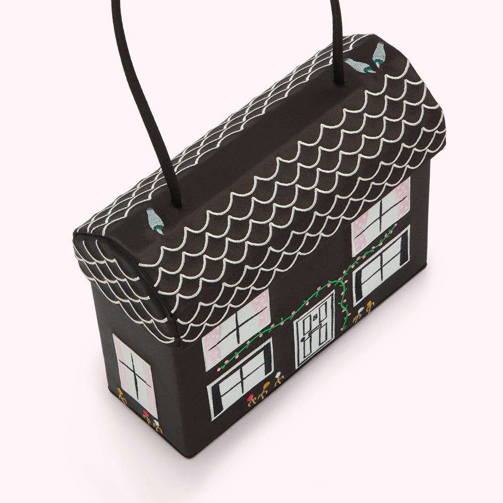 Black handbag shaped like a house with white scaled roof and pink windows, green vines, and small figures embroidered on the sides.
