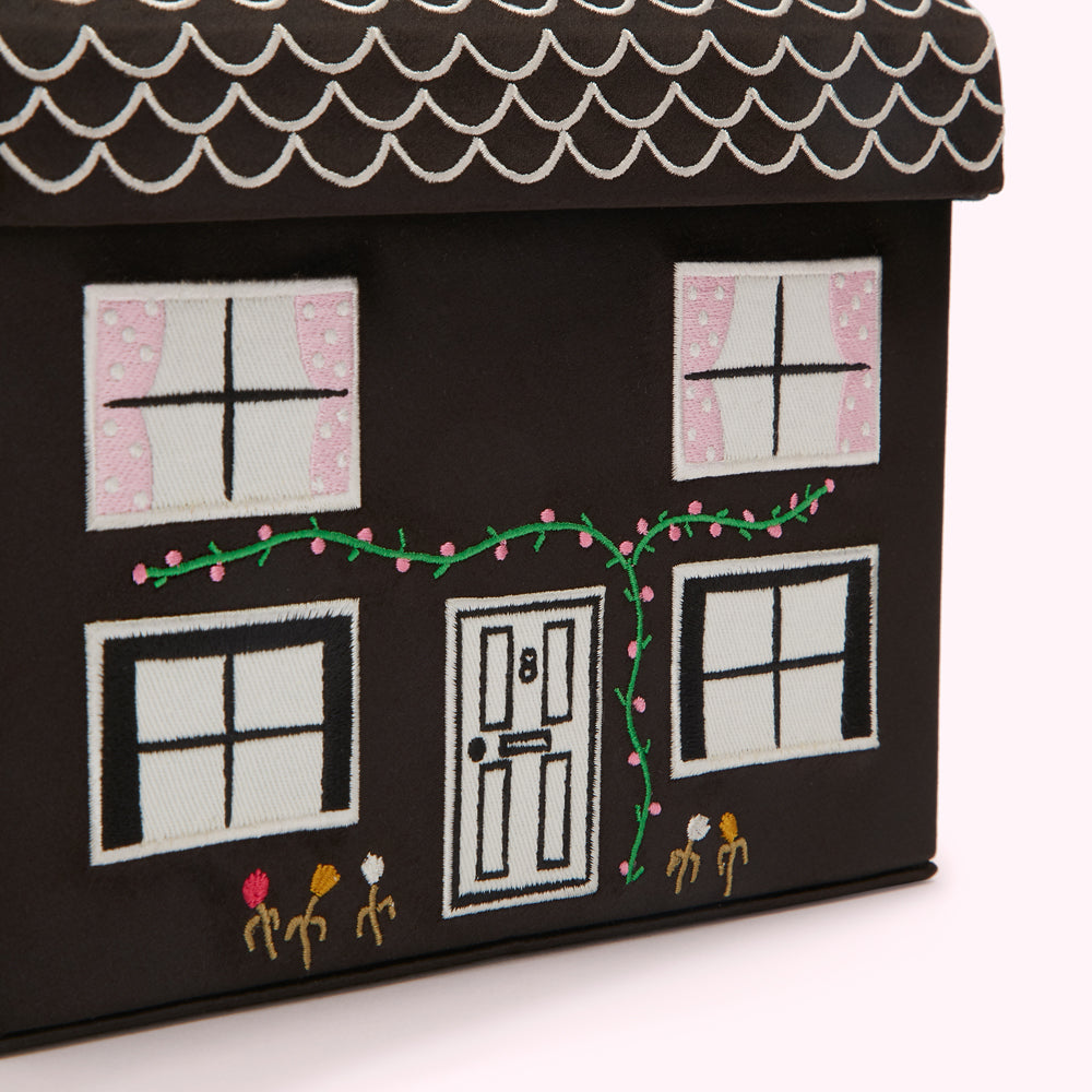Close-up of a dark brown house-shaped storage box with embroidered details, including windows, a door, and climbing vines.
