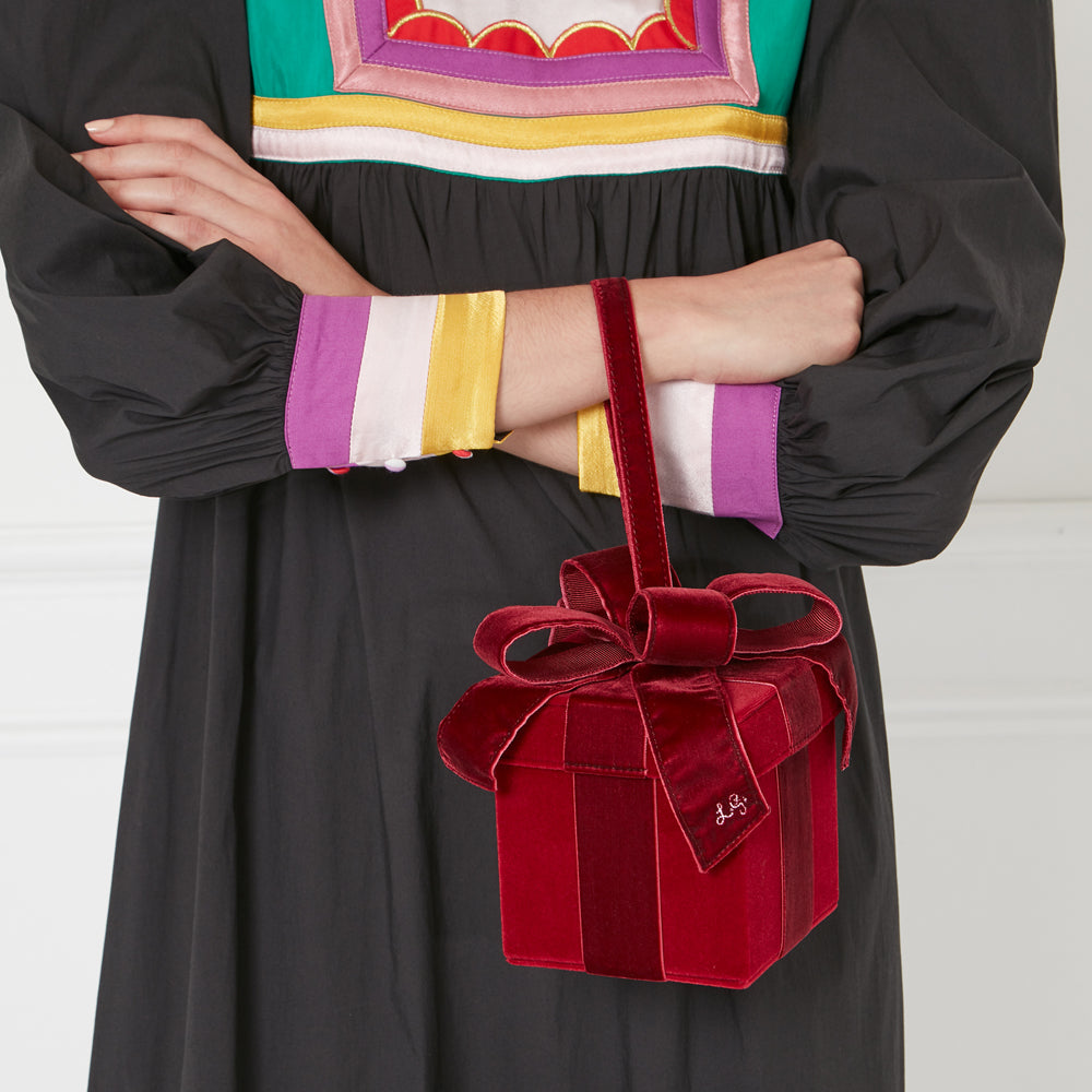 A woman in a black dress with colorful details holds a red velvet gift box-shaped handbag.
