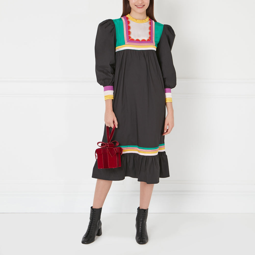 Woman in black midi dress with colorful trim and red gift bag, against white background.
