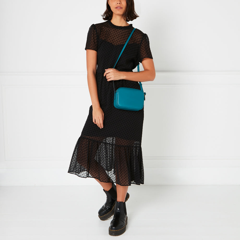 A woman in a black polka dot dress carries a teal crossbody bag.
