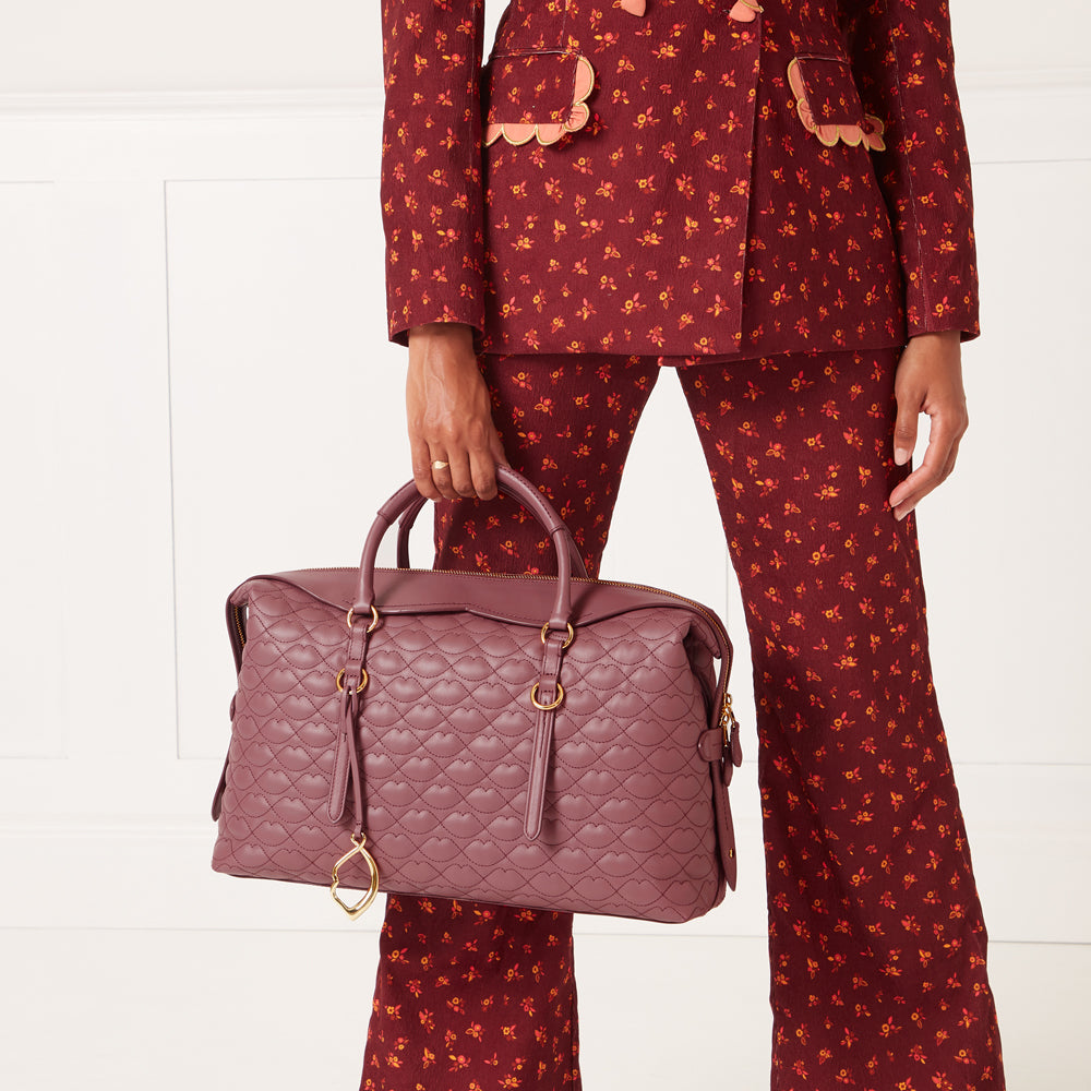A mauve quilted handbag with gold-tone hardware, held by a person wearing a burgundy floral pantsuit against a white background.
