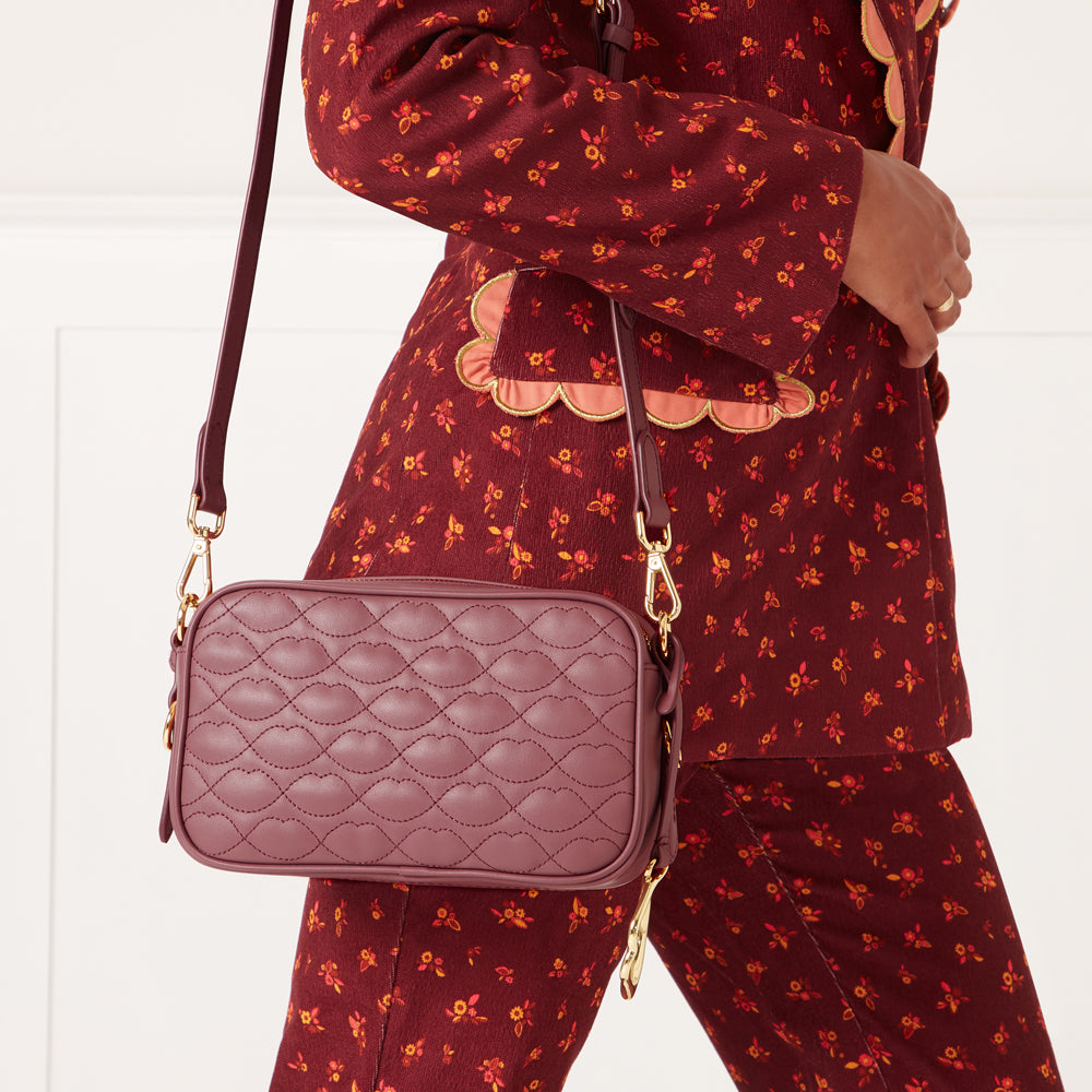 A mauve quilted crossbody bag with gold hardware hangs from the shoulder of a woman wearing a burgundy floral pantsuit.
