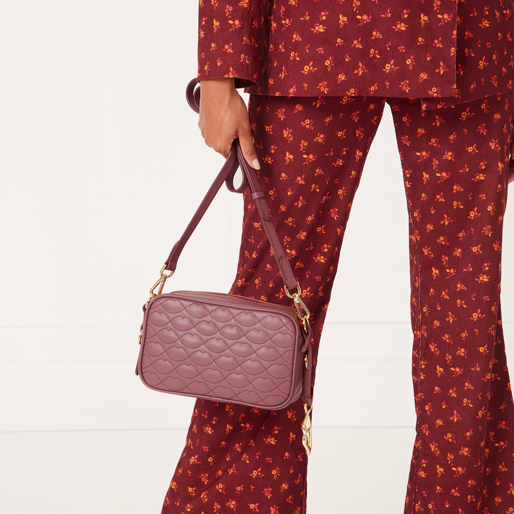 A maroon quilted crossbody bag with gold hardware is held by a person wearing a maroon floral pantsuit against a white background.
