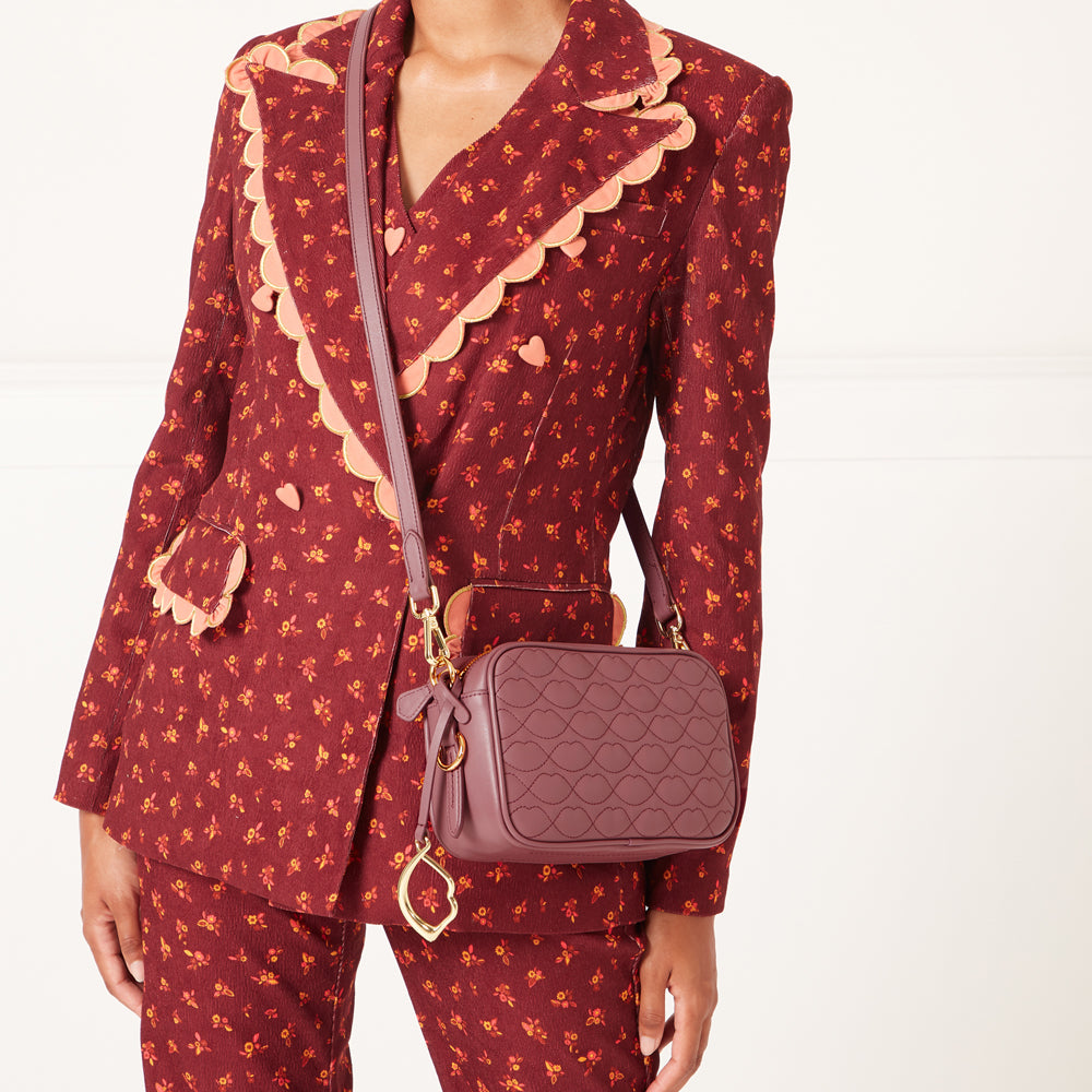 A maroon quilted crossbody bag with gold hardware hangs on a person wearing a maroon floral-print suit jacket.