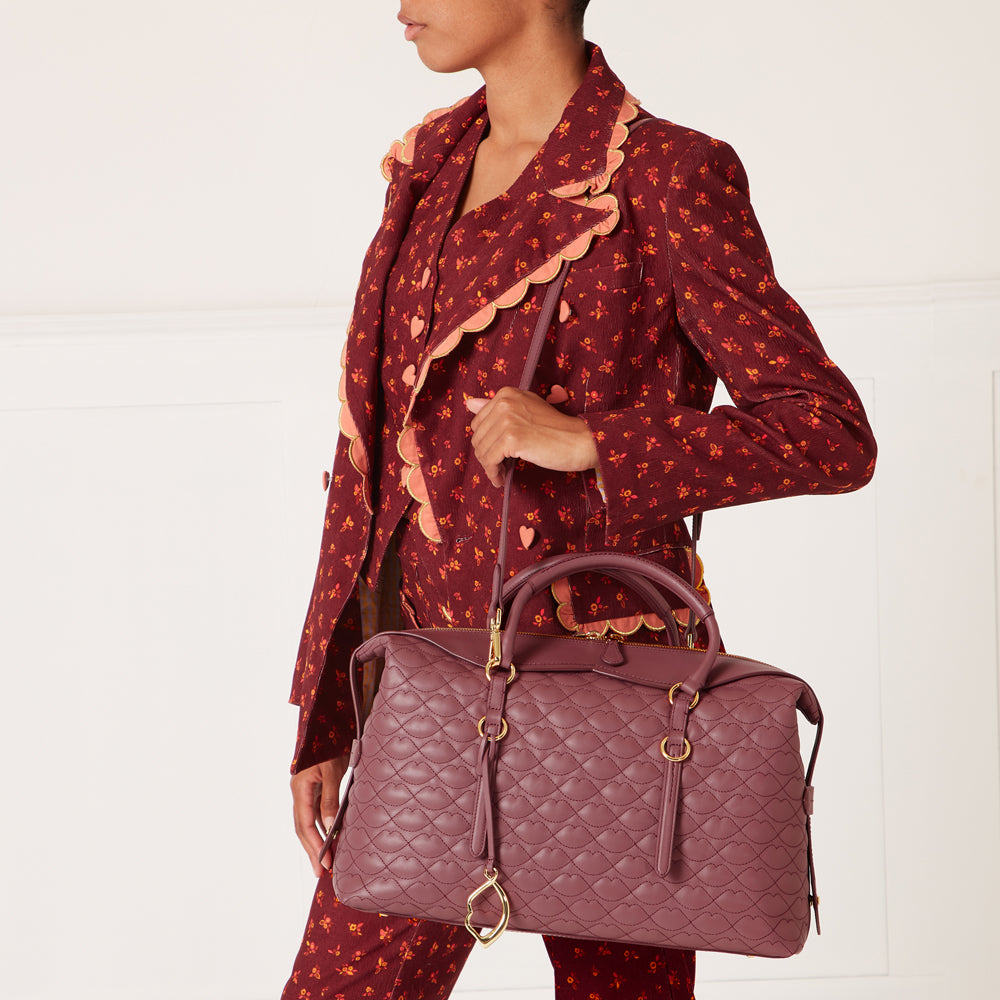 A mauve quilted handbag with gold accents hangs from the shoulder of a person wearing a maroon floral print jacket and matching pants against a white background.
