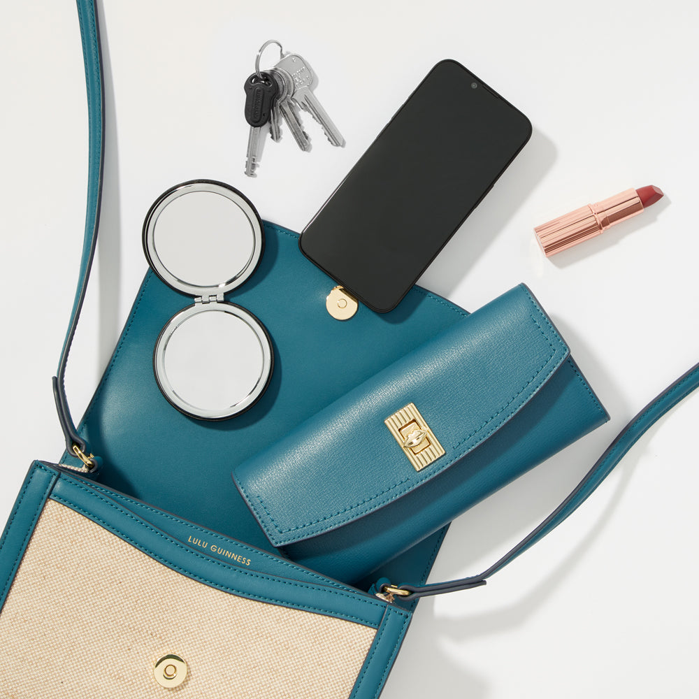A teal Lulu Guinness crossbody bag open to reveal its contents: a compact mirror, keys, smartphone, wallet, and lipstick.
