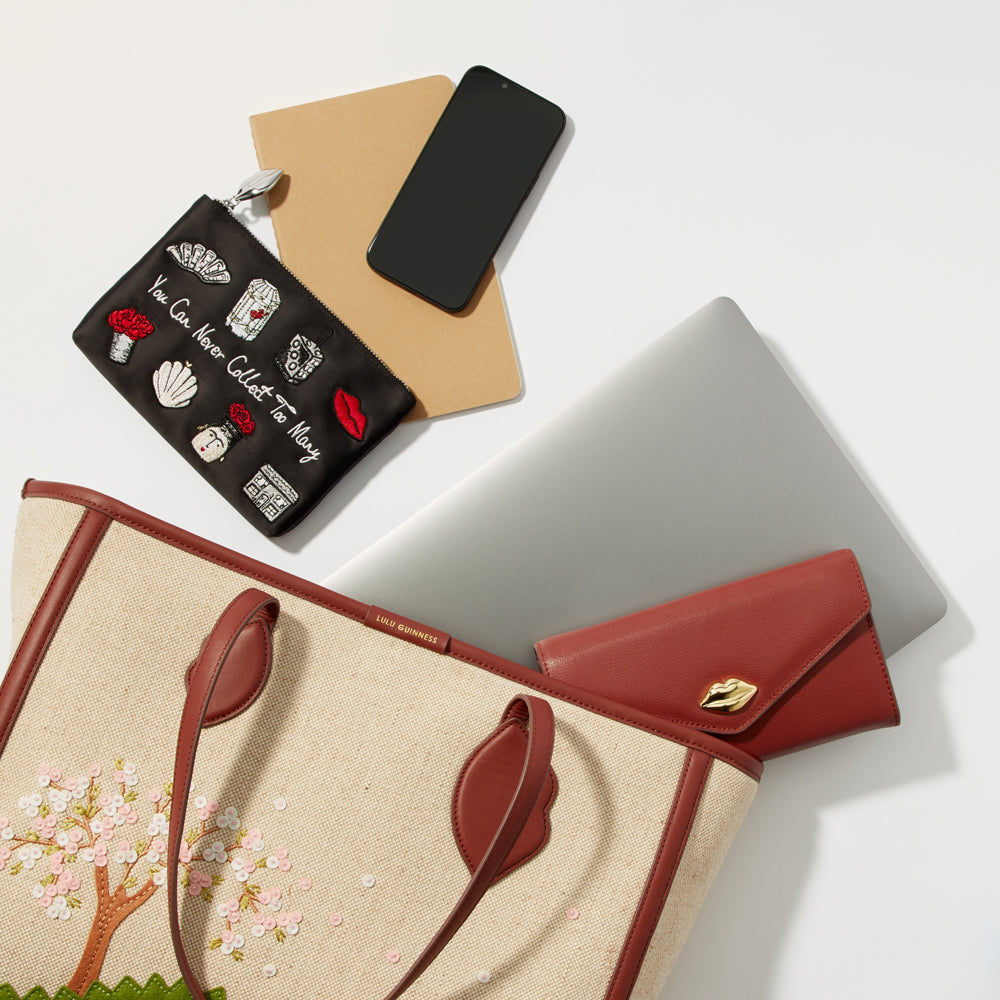 A beige tote bag with embroidered cherry blossoms, a black embroidered pouch, a brown notebook, a silver laptop, a red wallet, and a black smartphone, all arranged on a white background.