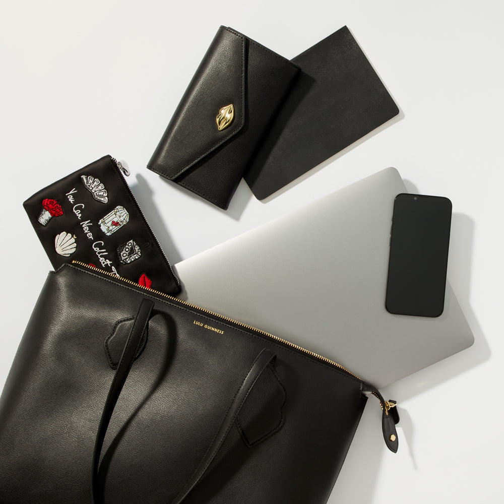 A black tote bag on a white background, open to reveal a laptop, phone, notebook and several smaller purses.