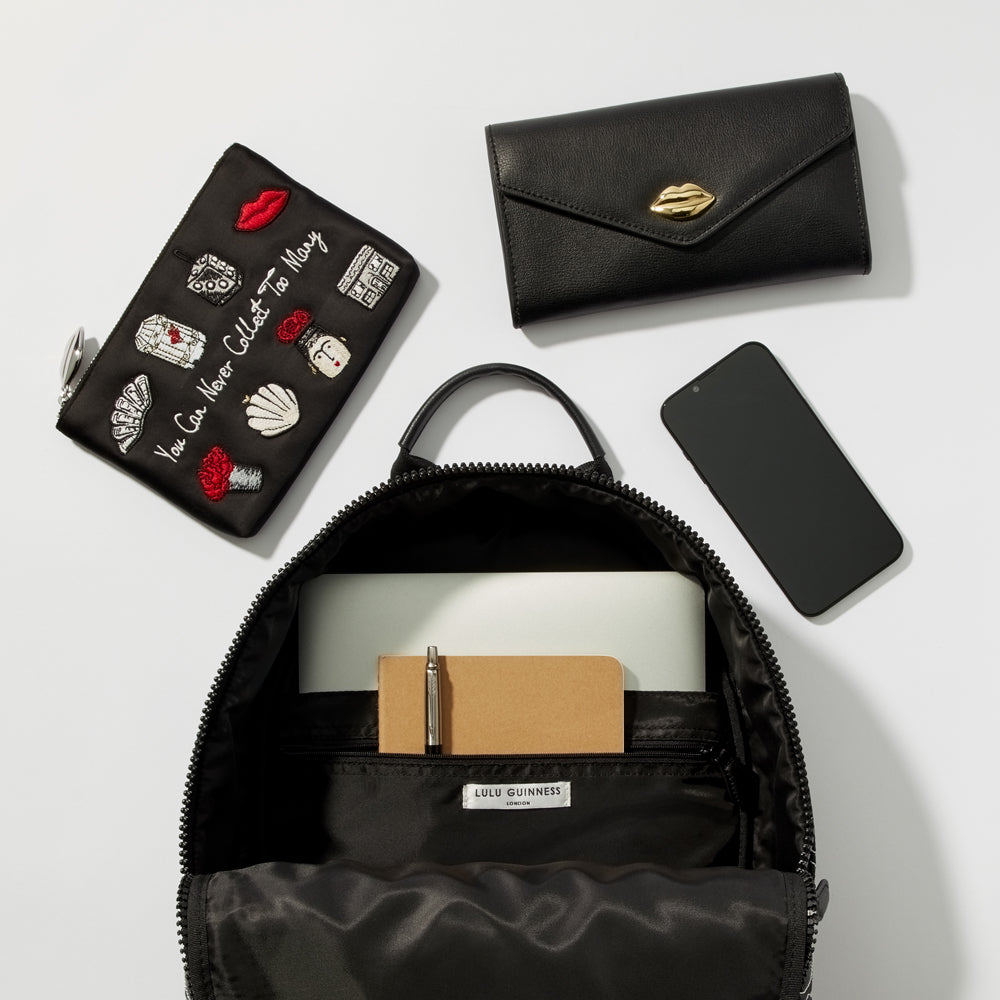 A black Lulu Guinness backpack is open to reveal its interior, which contains a laptop, notebook, and pen. A black leather wallet with a gold lip detail and a black cosmetic bag with embroidered details are also shown. A black smartphone is visible. The background is white.
