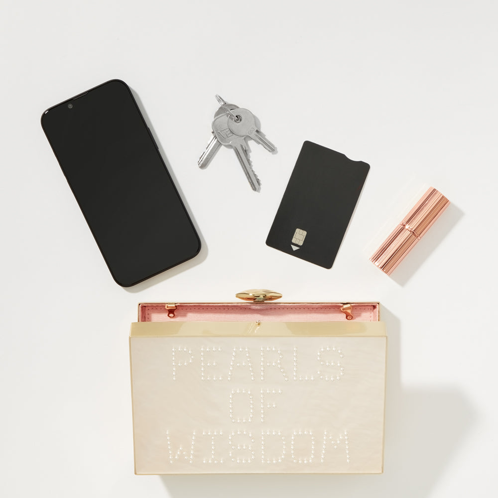 Flatlay of a smartphone, keys, credit card, lipstick, and a clutch purse that says "Pearls of Wisdom".
