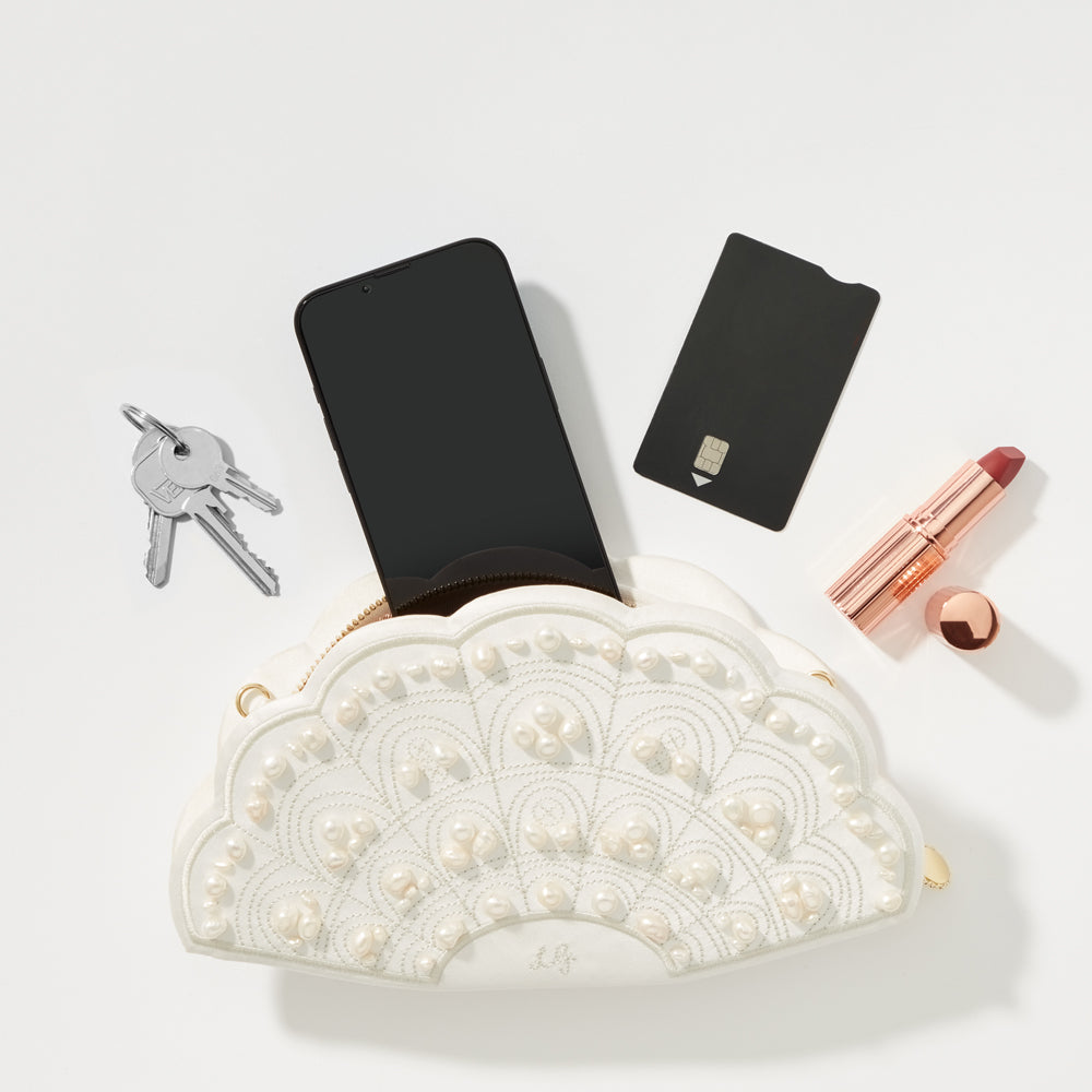 A white pearl-embellished fan-shaped clutch containing a smartphone, keys, and a credit card, with lipstick and its cap beside it, on a white background.
