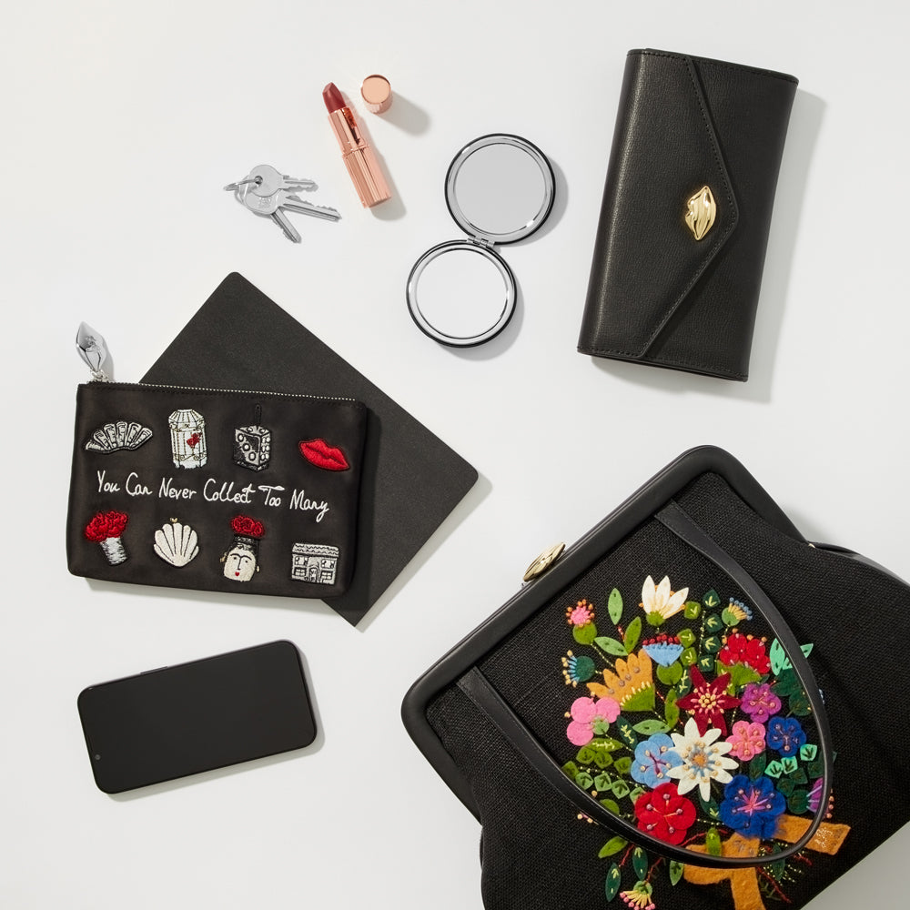 Flatlay of black accessories on a white background including a black handbag with embroidered flowers, a black wallet, a black make-up pouch with embroidered details, a compact mirror, lipstick, keys, and a black smartphone.