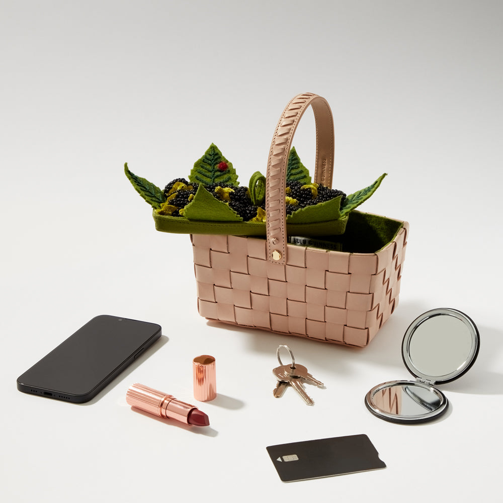 A pale pink woven basket containing a felt blackberry design sits on a white background, next to a smartphone, lipstick, keys and a compact mirror.
