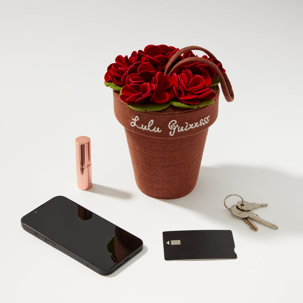 A brown fabric Lulu Guinness flower pot handbag with red flowers, next to a smartphone, lipstick, and keys.