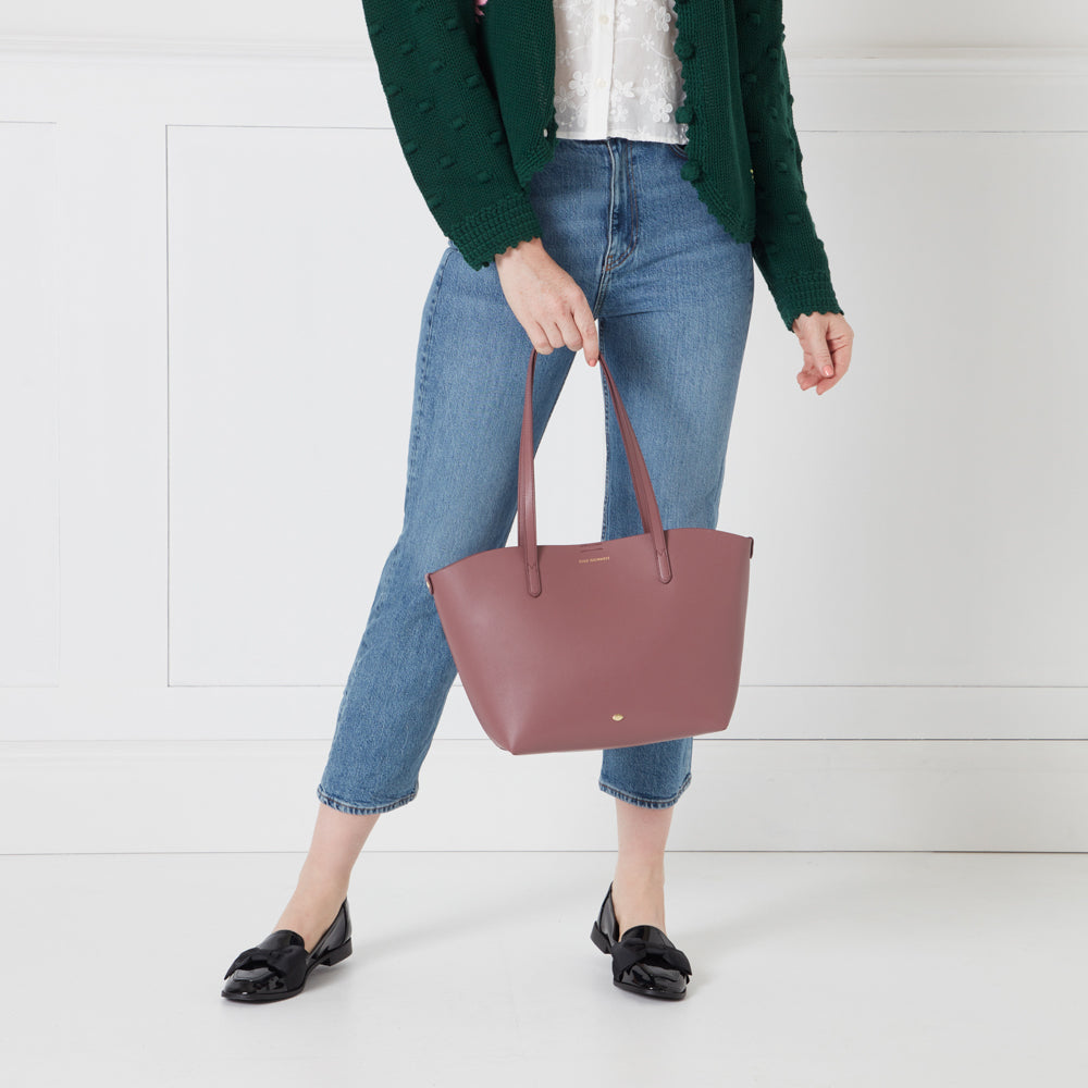 A woman in a green cardigan and jeans holds a dusty rose tote bag against a white background.