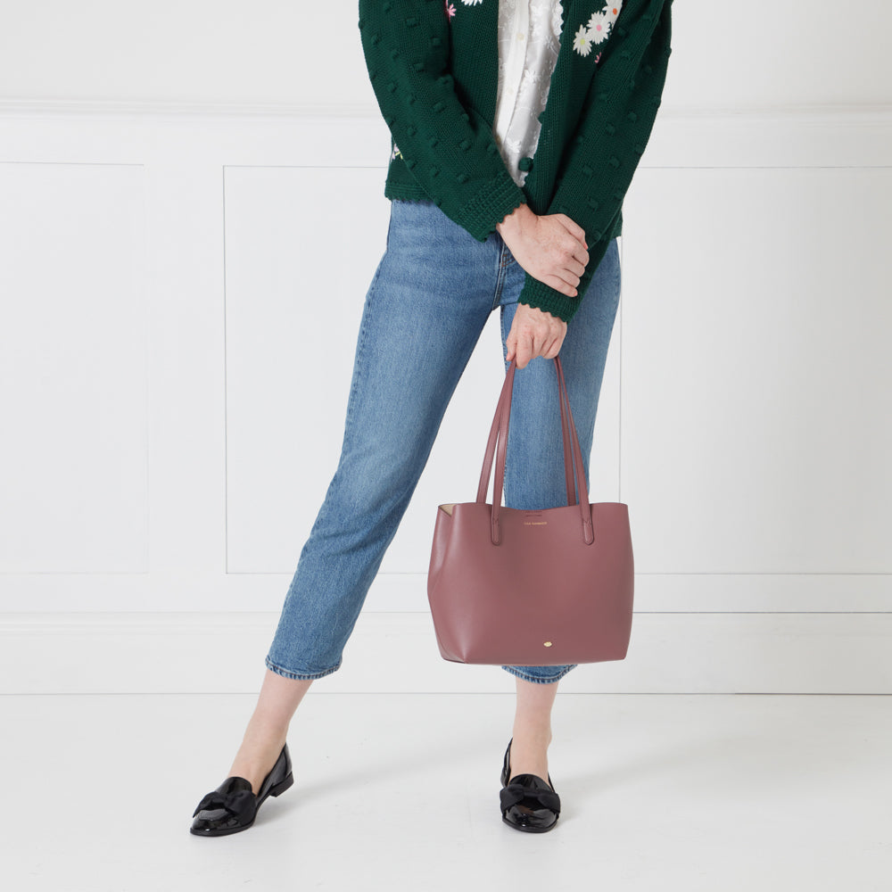 A woman in blue jeans and a green cardigan holds a dusty rose tote bag against a white background.
