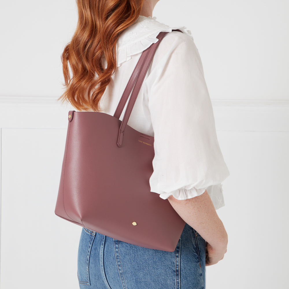 A woman with long, wavy auburn hair is shown from the back, wearing a white blouse and jeans. She carries a mauve-coloured Lulu Guinness tote bag.
