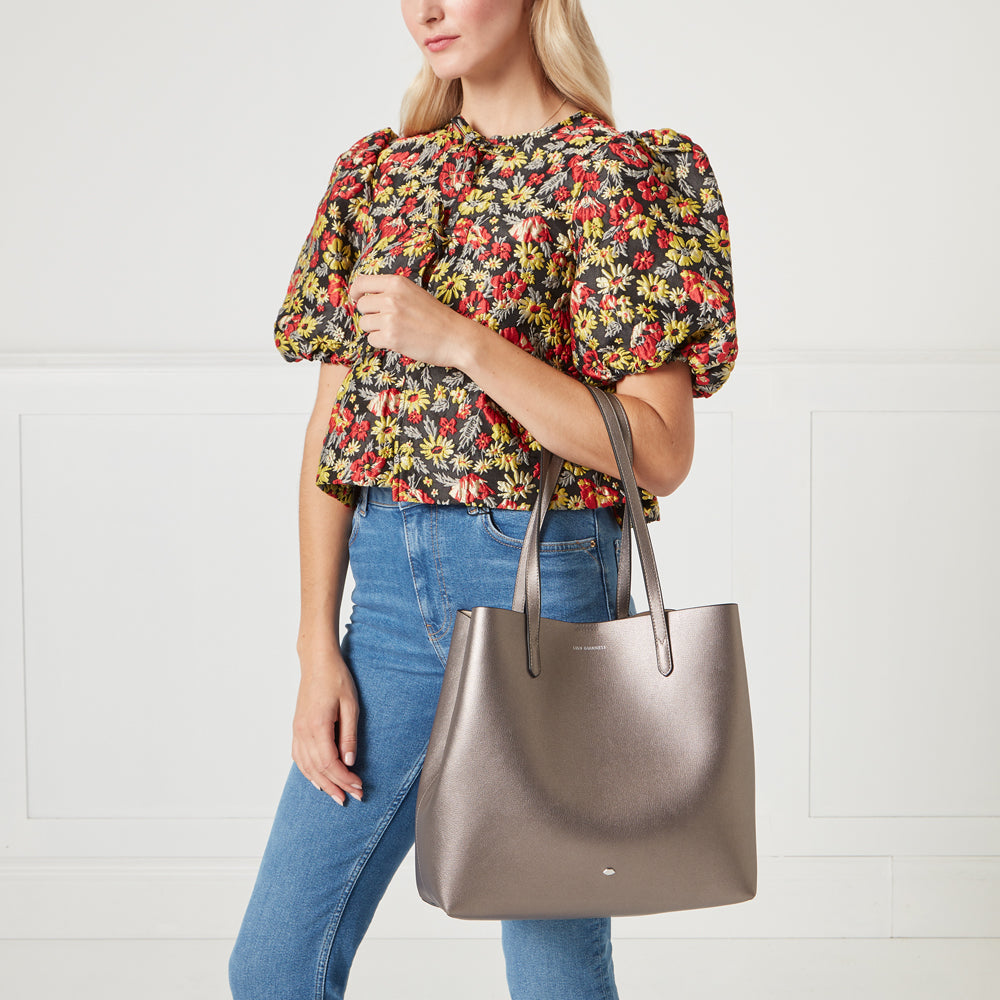 A woman in a floral top and jeans holds a metallic gunmetal grey tote bag.

