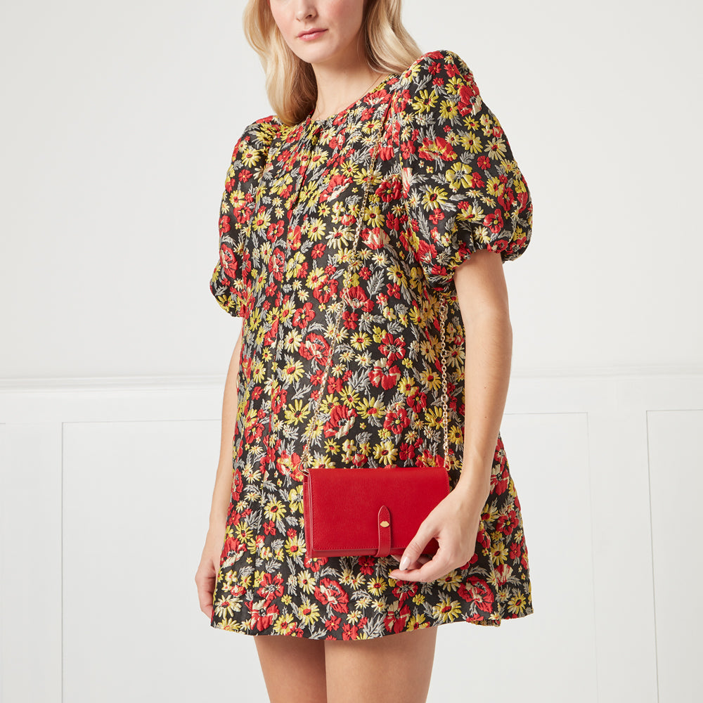 A woman in a floral dress holds a red crossbody wallet.
