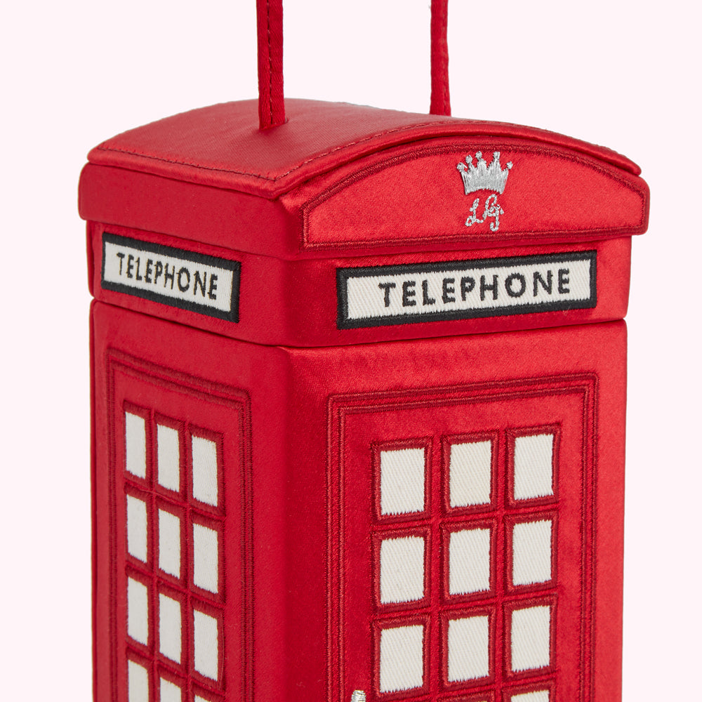 Close-up of a red satin handbag shaped like a London telephone booth, featuring embroidered details and a crown emblem.
