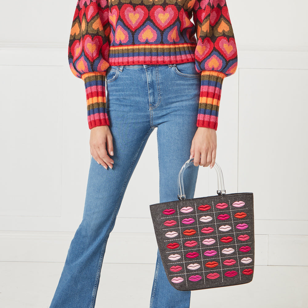 A woman in blue jeans and a rainbow heart sweater holds a grey felt tote bag with embroidered lips. The background is white.
