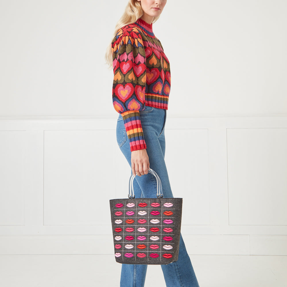 Woman in colourful heart sweater holding grey felt tote bag with embroidered lips against white background.
