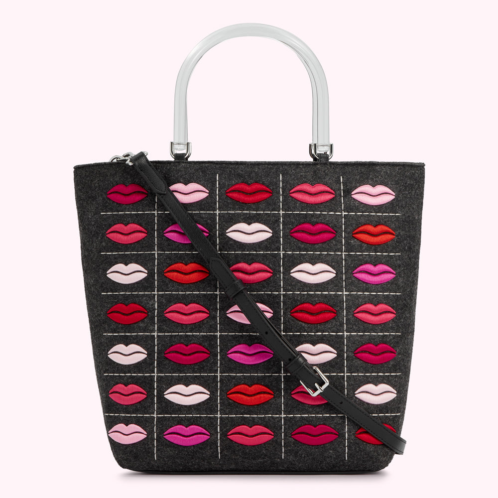 Gray felt tote bag with clear acrylic handles and a black crossbody strap, adorned with a grid pattern of embroidered lips in various shades of pink and red.
