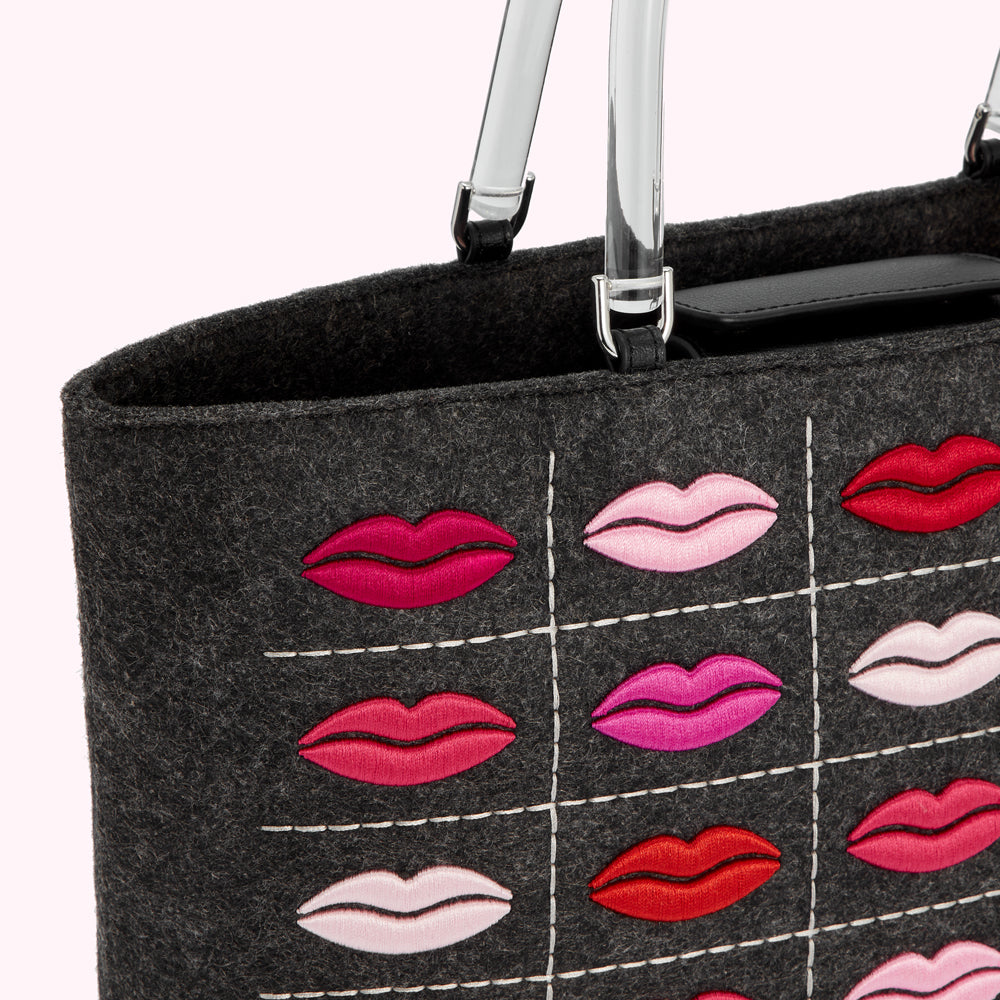 Close-up of a dark grey felt tote bag with clear acrylic handles and a pattern of embroidered lips in various shades of pink and red.