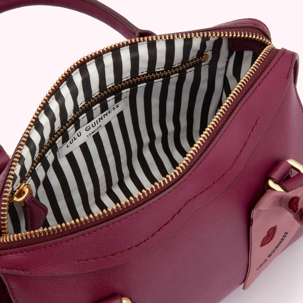 Close-up of a burgundy Lulu Guinness handbag open, showcasing its black and white striped interior and gold zipper. A small red lip detail is visible on the exterior.
