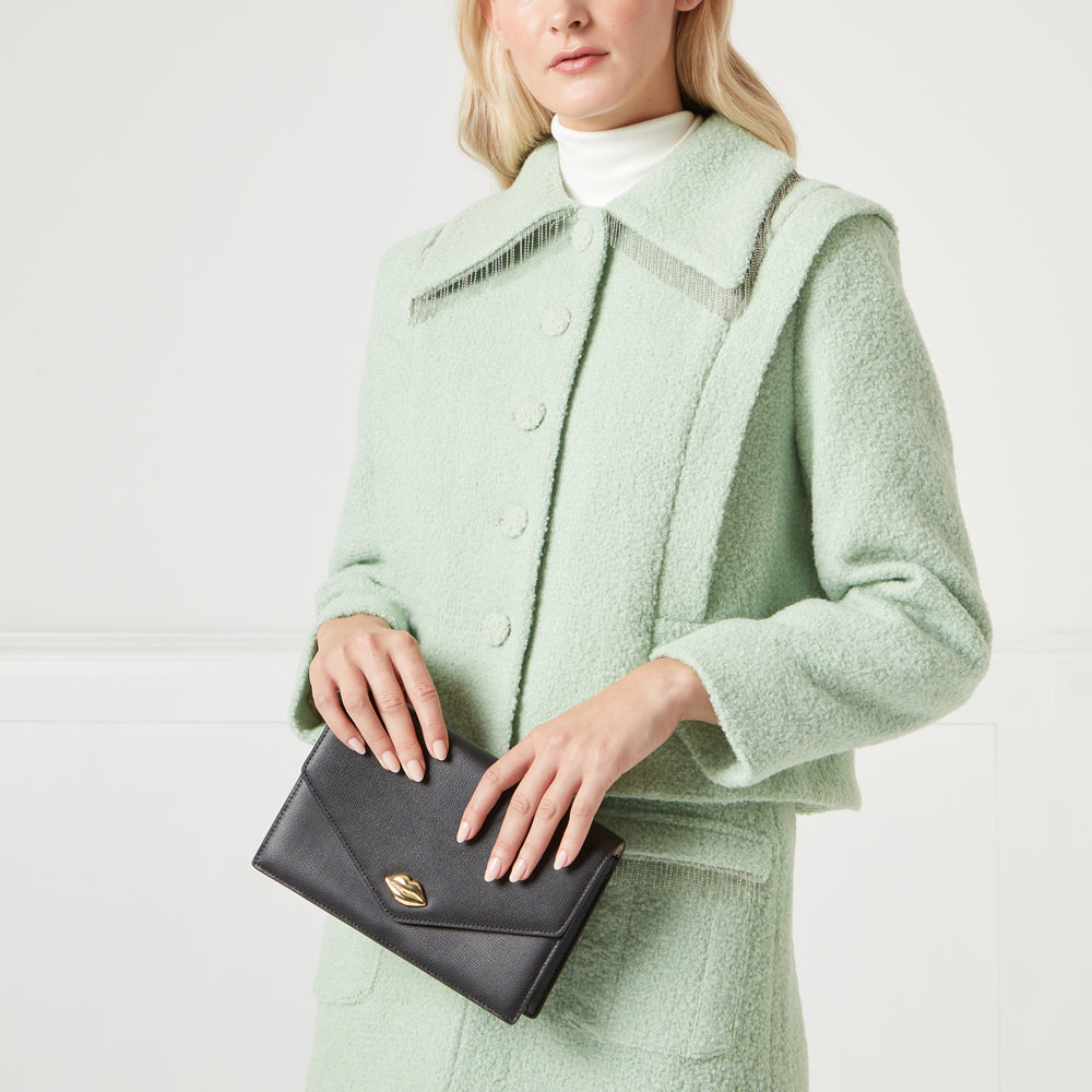 A woman in a pale green textured jacket holds a black clutch purse with a gold lip-shaped detail.