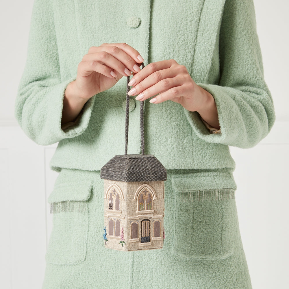 A woman in a mint green coat holds up a small, beige house-shaped handbag with dark grey trim and embroidered details.