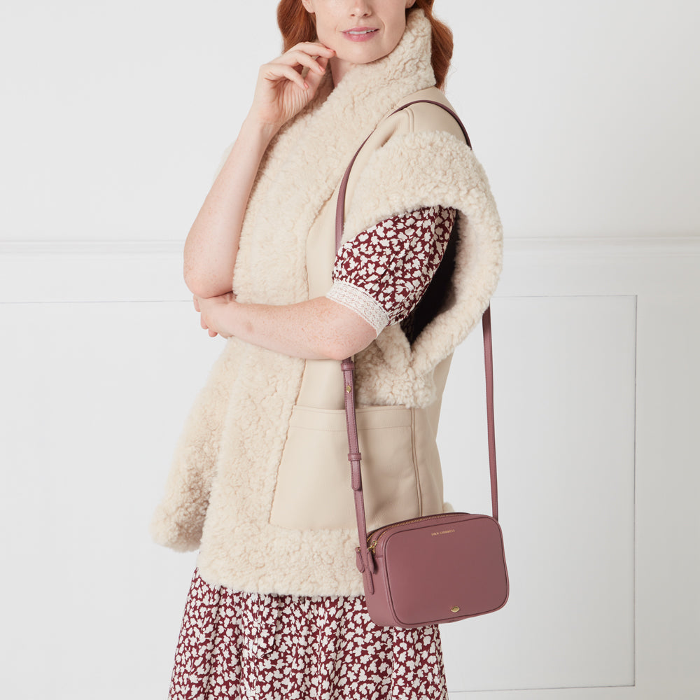 A woman wearing a cream-colored sheepskin vest and a burgundy floral dress carries a dusty rose-coloured crossbody bag.
