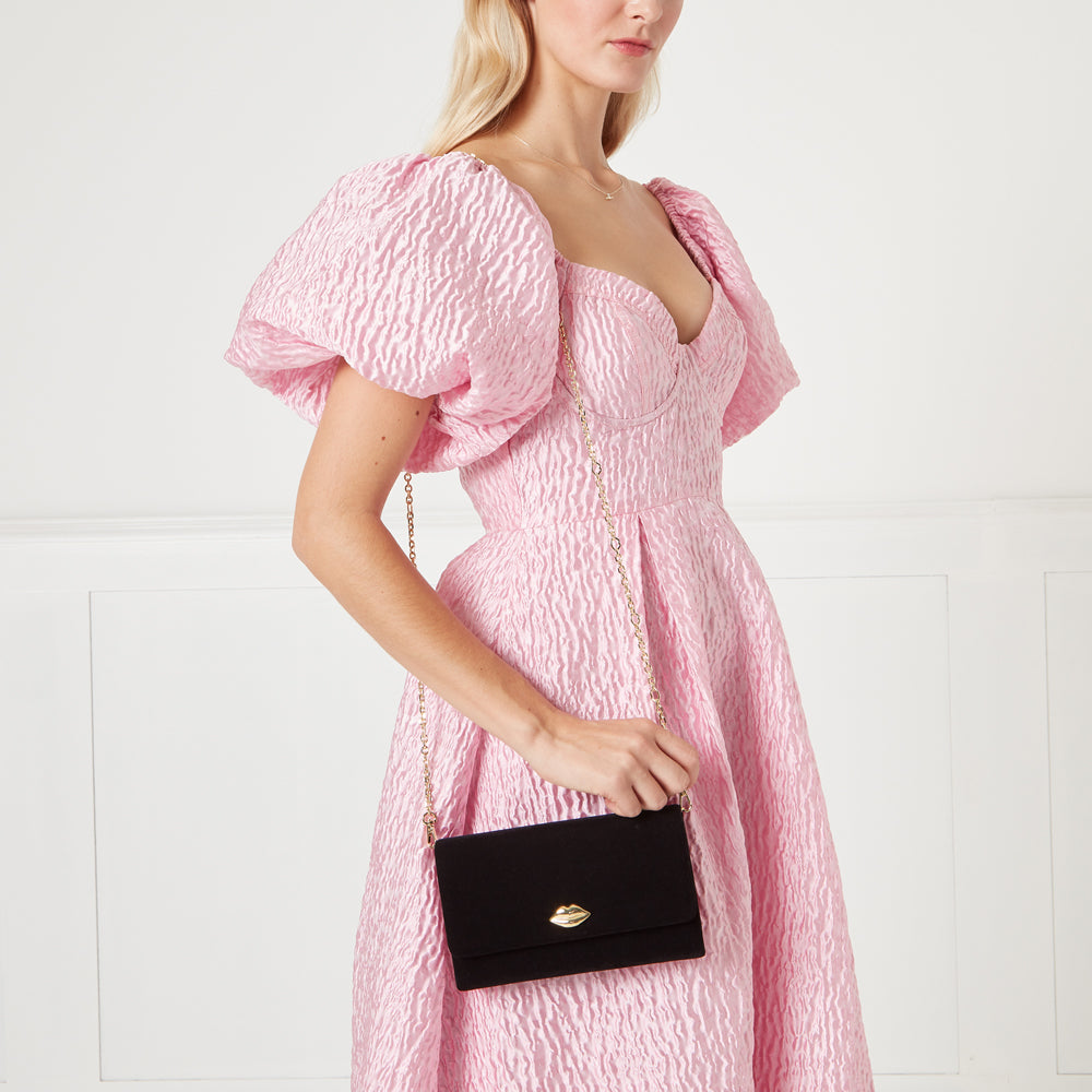 Woman in pink dress holding black velvet crossbody bag with gold lip clasp. Background is off-white.
