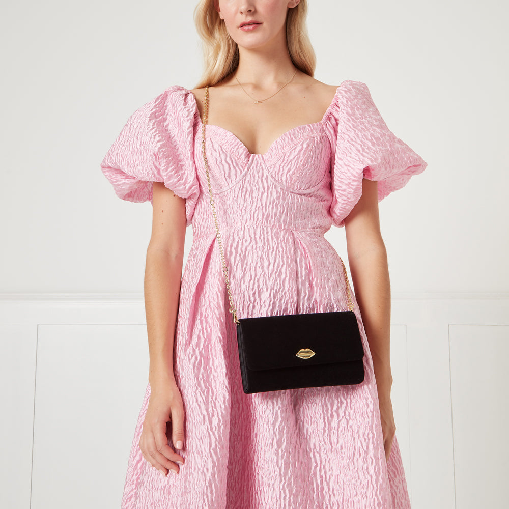 Woman in pink dress holding black velvet crossbody bag with gold lip detail.
