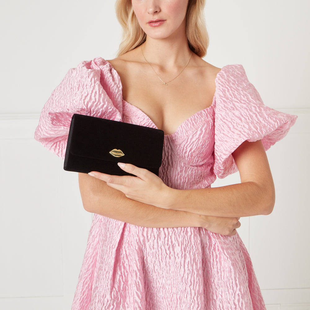 Woman in pink dress holding black velvet clutch with gold lip detail.
