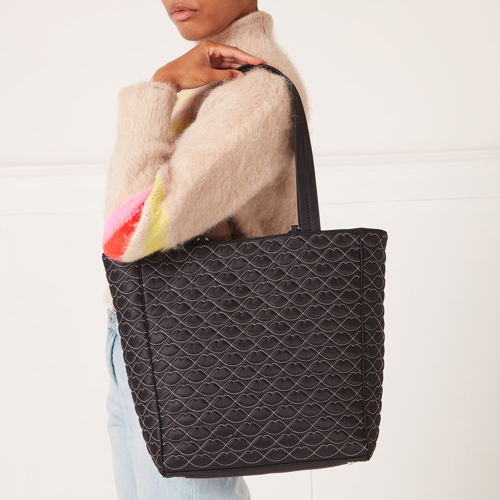 Black quilted tote bag with a lip print pattern, held by a person wearing a light beige sweater.
