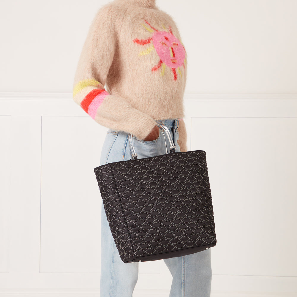 Woman in light beige fuzzy sweater and light blue jeans holding a large black quilted tote bag with clear handles. The bag features a repeating lip print. The background is white.
