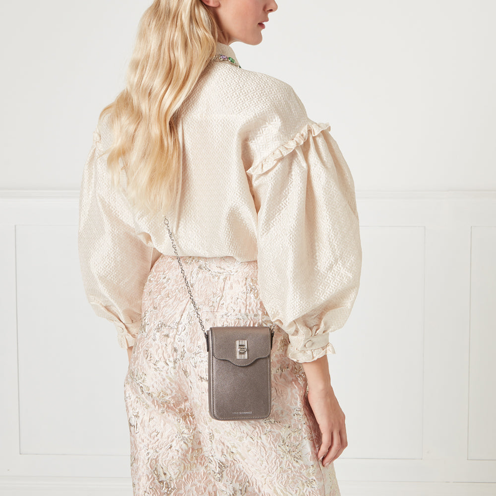 Woman wearing a beige blouse and a pink brocade skirt, carrying a gunmetal grey crossbody phone bag.
