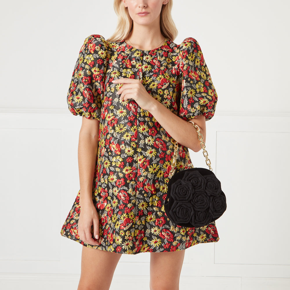 Woman in floral dress holding a black rose-shaped handbag with a gold chain strap against a white background.
