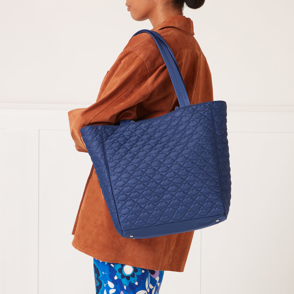 A navy blue quilted tote bag with a lip print pattern, being carried by a person wearing a brown shirt and blue pants.
