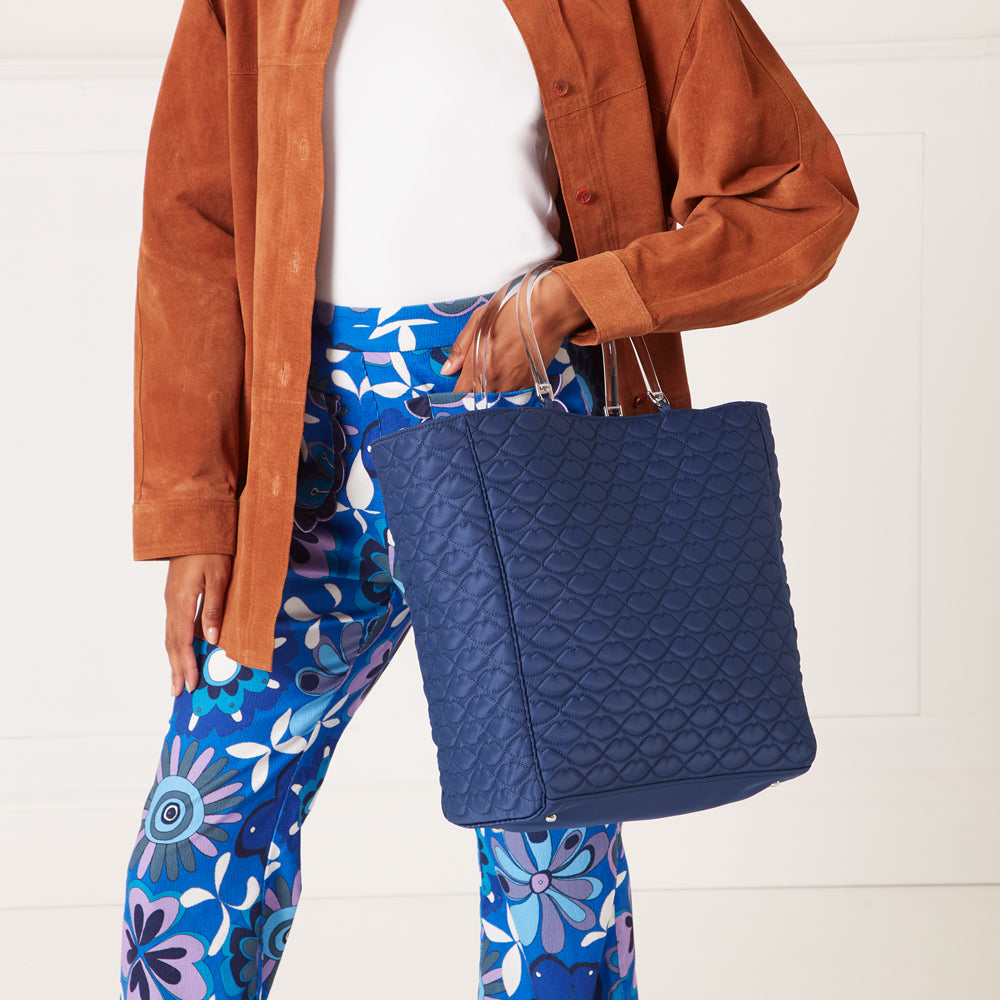 A navy blue quilted tote bag with clear handles is held by a person wearing a brown suede jacket and blue floral pants against a white background.
