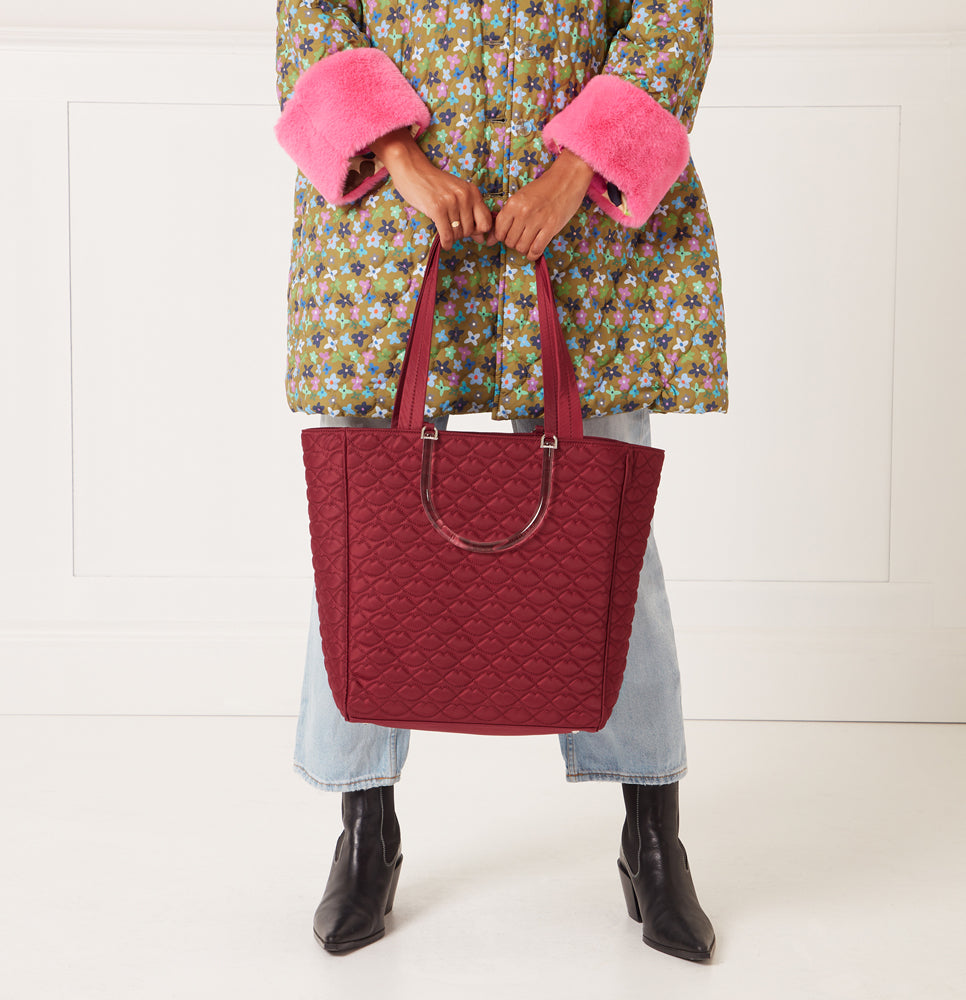 A woman in a floral quilted coat and light blue jeans holds a large burgundy quilted tote bag with translucent handles.