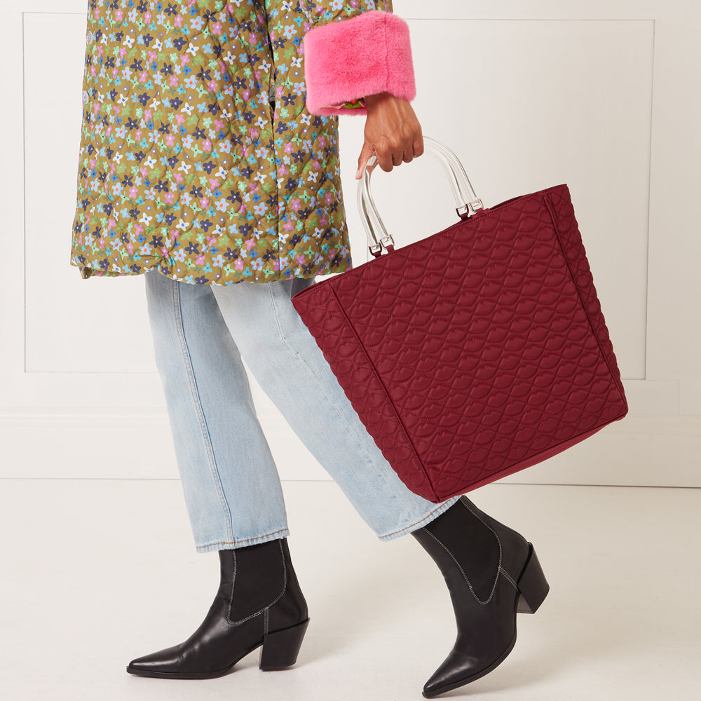 A person in light blue jeans, black boots, and a floral quilted jacket carries a large quilted burgundy tote bag with clear handles against a white background.