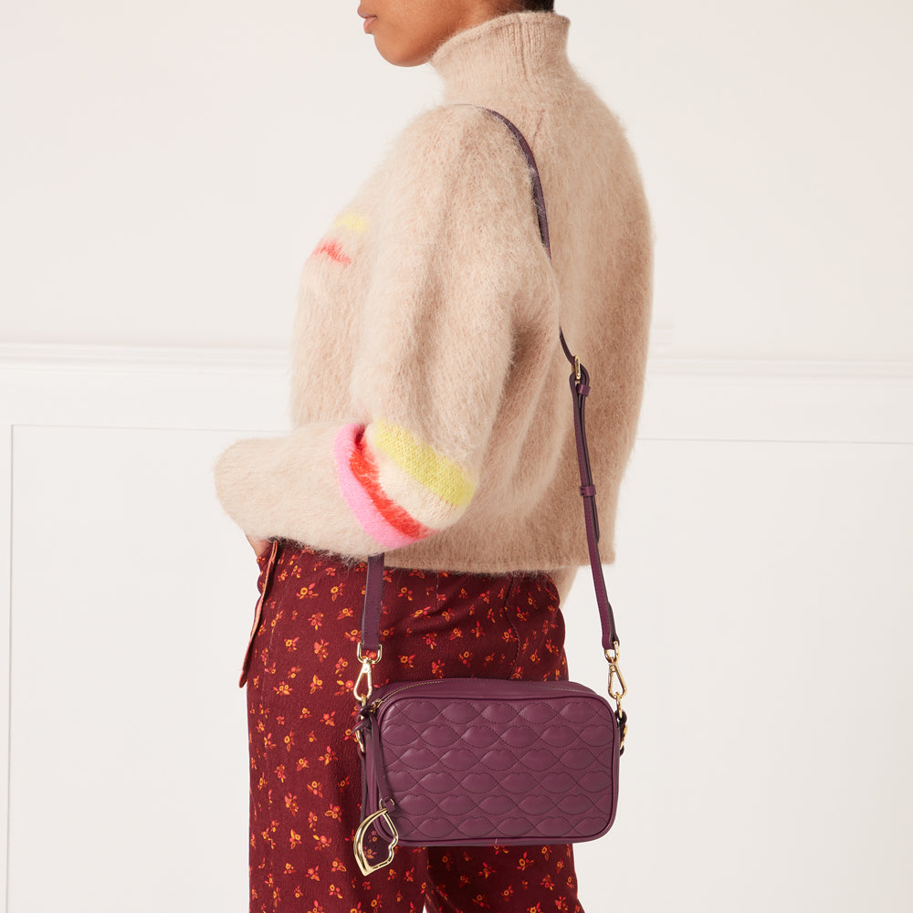 A woman in a beige fuzzy sweater and maroon floral pants carries a quilted purple crossbody bag.
