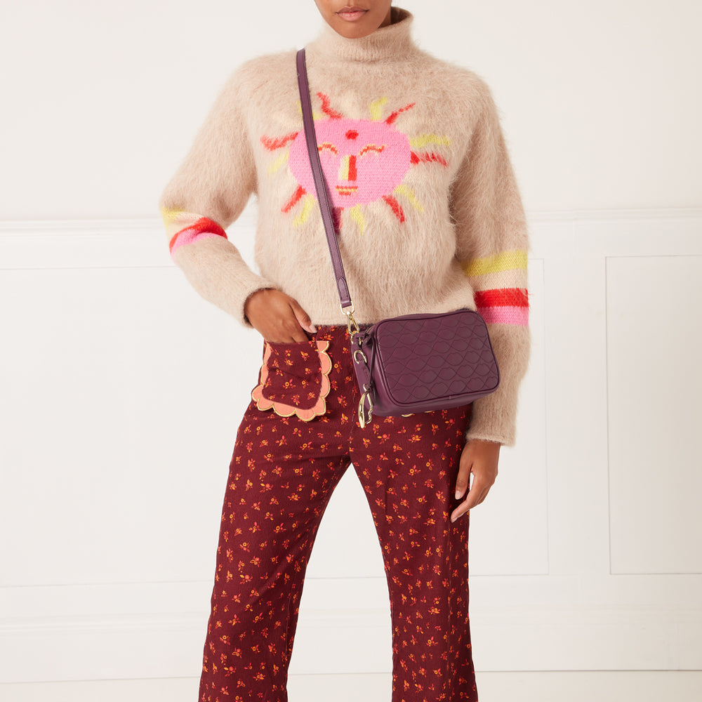 A woman models a deep purple quilted crossbody bag against a white background. She is wearing a fluffy beige sweater with a pink sun design and burgundy floral pants.
